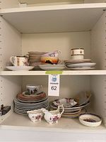 Photo of shelf with assorted vintage china including stacked saucers, teacups, cream and sugar sets, and decor plates with floral designs.