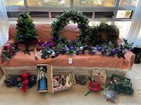 Wide view of lot showing multiple Christmas trees, wreaths, garlands, figurines and boxed angel topper in original packaging arranged on a couch covered with a pink polka dot cloth.
