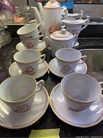 Photo of 6 cups and saucers arranged with coffee pot, creamer, and sugar bowl in background showing ship motif and gold accents
