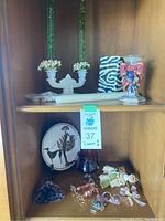 Full frontal image showing multiple decorative items arranged on two wooden shelves including green glass candle holders, zebra print vase, pink ribbon fruit motif bowl, figurines, candles and decorative plate.