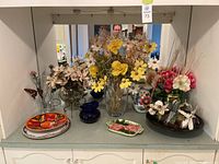 Photo of multiple artificial flowers arrangements in cream, yellow, pink alongside various glass and ceramic vases, decorative plates, and a blue glass vase.