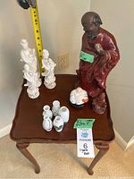 Five figurines and five small vases arranged on a wooden side table, with measuring tape showing height of larger figurine around 10 inches. Large figurine is a man in red robe and is chipped on the hand. Several white porcelain figurines and small vases with floral detail.