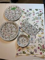 Top view of 2 ceramic plates, 1 bowl, rectangular trivet, and 6 floral placemats arranged on table, showing the entire set and floral pattern