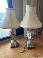 Pair of vintage ceramic table lamps with floral designs, beige fabric lampshades, and cords visible on hardwood floor near glass door and curtains.