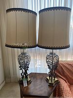 Two matching glass table lamps with beige fabric shades placed on a wooden table in front of a sheer curtain. Lamps have ornate black metal scrollwork inside clear glass bodies.