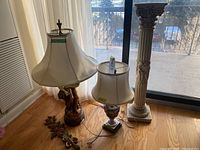 Wide view of two table lamps with fabric shades and one Corinthian-style pedestal column placed on wooden floor near window.