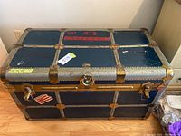Front view of the blue metal steamer trunk showing brass hardware and latch with padlock clasp. Visible scratches and marks on surface with a label sticker on front.