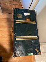 Top and front views of the green metal steamer trunk showing cream stripes and metal corner protectors, with visible scratches and travel labels.