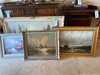 Four vintage framed paintings and prints leaning against furniture, showing different frame styles and sizes.