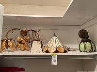 Set of four vintage light fixtures displayed on a shelf, showing gold metal leaf design fixtures, a fabric plug-in lamp with beaded fringe, and a green stained glass fixture.