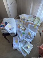 Overview of assortment of vintage linens and tablecloths displayed on two small tables, showing different items stacked and laid out with some having sticky notes indicating quantities.