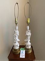 Pair of vintage oriental porcelain table lamps on a wooden table, showing the full lamp with white porcelain figure bases and brass bottom, topped with lamp fittings sans shades.