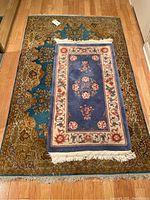 Top view of two stacked rugs on a wooden floor, showing the larger teal rug with floral and geometric patterns beneath a smaller blue and cream rug with floral border and fringes.