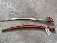 Full view of the sabre and matching red fabric sheath with brass fittings, placed on a light carpet background.