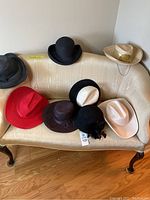 Eight women's hats displayed on a cream-colored loveseat including different styles like cloche, cowboy, bowler, and wide-brim hats in various colors.