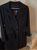 Black coat hanging showing front, single-breasted with belt, long sleeves with button detail.