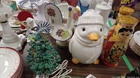 Photo showing ceramic Christmas tree with lights, two ceramic roosters with floral patterns, penguin cookie jar, and Millennium nesting dolls grouped together, displayed on table with other ceramics.