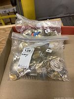 Two sealed plastic bags filled with assorted metal costume jewellery fasteners and parts placed in a cardboard tray.