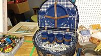 Blue wicker picnic basket open showing blue plastic plates, cups, and utensils for four, set inside with plaid lining