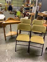 Photo showing solid wood corner table and four folding chairs with green fabric seats in a warehouse environment