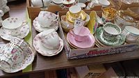 Photo showing multiple teacups and saucers of various patterns arranged in a box and on table, including pink, green, gold, and mixed floral designs.