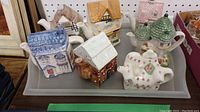 Tray containing six house-shaped ceramic teapots and one decorative chair-shaped ceramic piece with floral design.