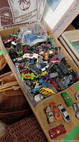 Box packed with many assorted toy cars including various models and colors, some wrapped in plastic bag