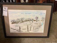 Framed watercolour painting showing a snow-covered rural landscape with trees, fence, and houses, mounted on neutral mat board and dark frame.