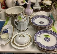 Photo showing Wedgwood Elgin plates stacked with purple and gold patterned rim, Mason's tureen bowl, child's tea set, ironstone teapot, and additional dinnerware items on table.