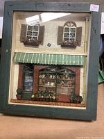 Photo of flower shop diorama with window shutters, fabric awning, and flower details in a distressed green frame.