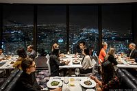 View of diners seated at tables inside a revolving restaurant with floor-to-ceiling windows overlooking a nighttime cityscape