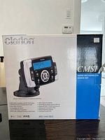 Photo of the Clarion Marine CMS1 source unit in its original packaging box against a plain background with a thermostat and light switch on the wall behind.