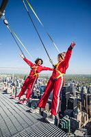 Two participants in red jumpsuits and harnesses walking hands-free on the edge of the CN Tower with city skyline below.