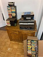Photo of three 8 track players stacked on a wooden cabinet with multiple stacks of 8 track tapes nearby.