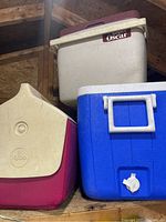 Three plastic coolers stacked in storage: one blue with spigot, one beige labeled 'Super Oscar', one cream and red Igloo cooler