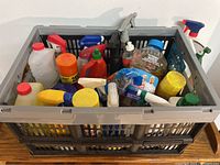 Plastic crate filled with assorted cleaning products and supplies including sprays, liquids, bottles with pump and screw caps.