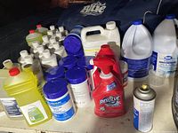 Photo shows a variety of cleaning products including multiple white bottles with blue caps and labeling, yellow bottles of concentrated neutral cleaner, a red spray bottle of Resolve, large blue and white bleach containers, and a metal aerosol canister.