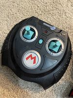 Remote control with dual joysticks and Mario logo on large central button, black with blue joysticks and power switch.
