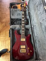 Red electric guitar inside grey plush-lined hardshell case with clip-on tuner and extra string pack