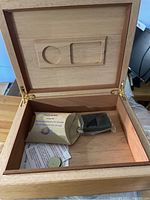Open wooden humidor box showing interior with humidity control device, small round humidity pack, and papers inside.