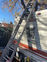 Two standing aluminum ladders leaning against a trailer with some fallen leaves and a man standing nearby.