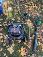 Yard Machines black push lawn mower, Weed Eater trimmer, and Weed Eater blower laid on grass with autumn leaves.