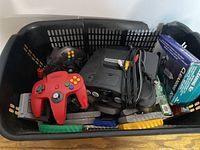 Top-down view of black basket containing console, controllers, cables, cleaning kit box, and cartridges.