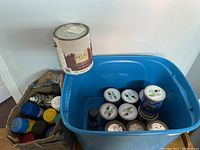 Overview photo showing blue plastic tote with multiple cans of wood stain and spray paint plus separate cardboard box with assorted spray paint cans and one white Relax paint can on top