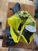 Paintball hoppers in a yellow plastic bag on wooden surface showing several black hoppers with clear lids.