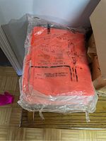 Photo showing bright orange raincoats sealed in plastic on stacked wooden chair or surface.