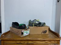 Photo of New Balance running shoe on its box and a black toe guard on its box, set on wooden surface.