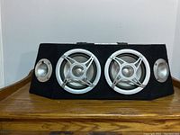 Front view of black fabric-covered trapezoidal speaker enclosure with two silver woofers and two smaller tweeters.