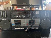 Front view of Toshiba black stereo with radio tuner, two cassette decks, and speakers, showing control knobs and power cord neatly coiled on top.