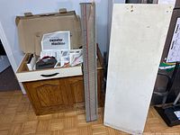 Photo showing Sweater Machine knitting machine unit in box alongside long needle board and white table board leaning against cabinet.